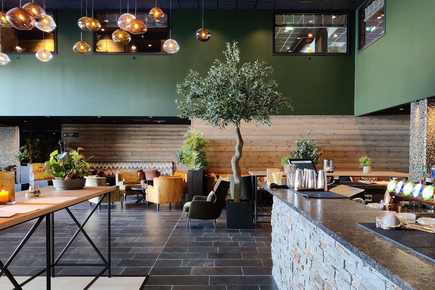 Restaurant interiour with slate floor, stone bar counter, a warm wooden panel on the wall with green shielding and many warm yellow hanging lamps. An 