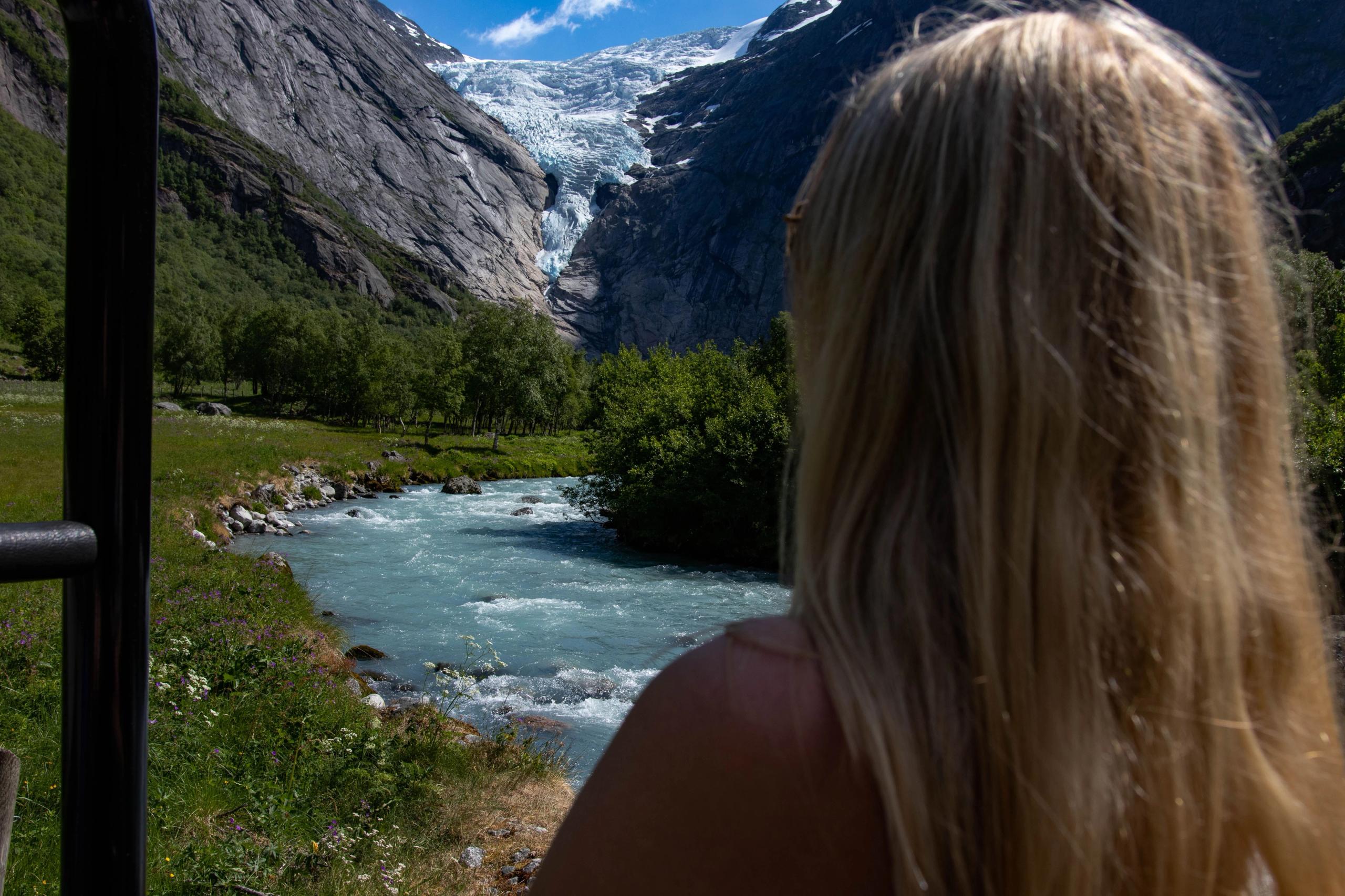 Rear view of a blonde person looking over a turquoise river toward a glacier between steep rocky mountains..