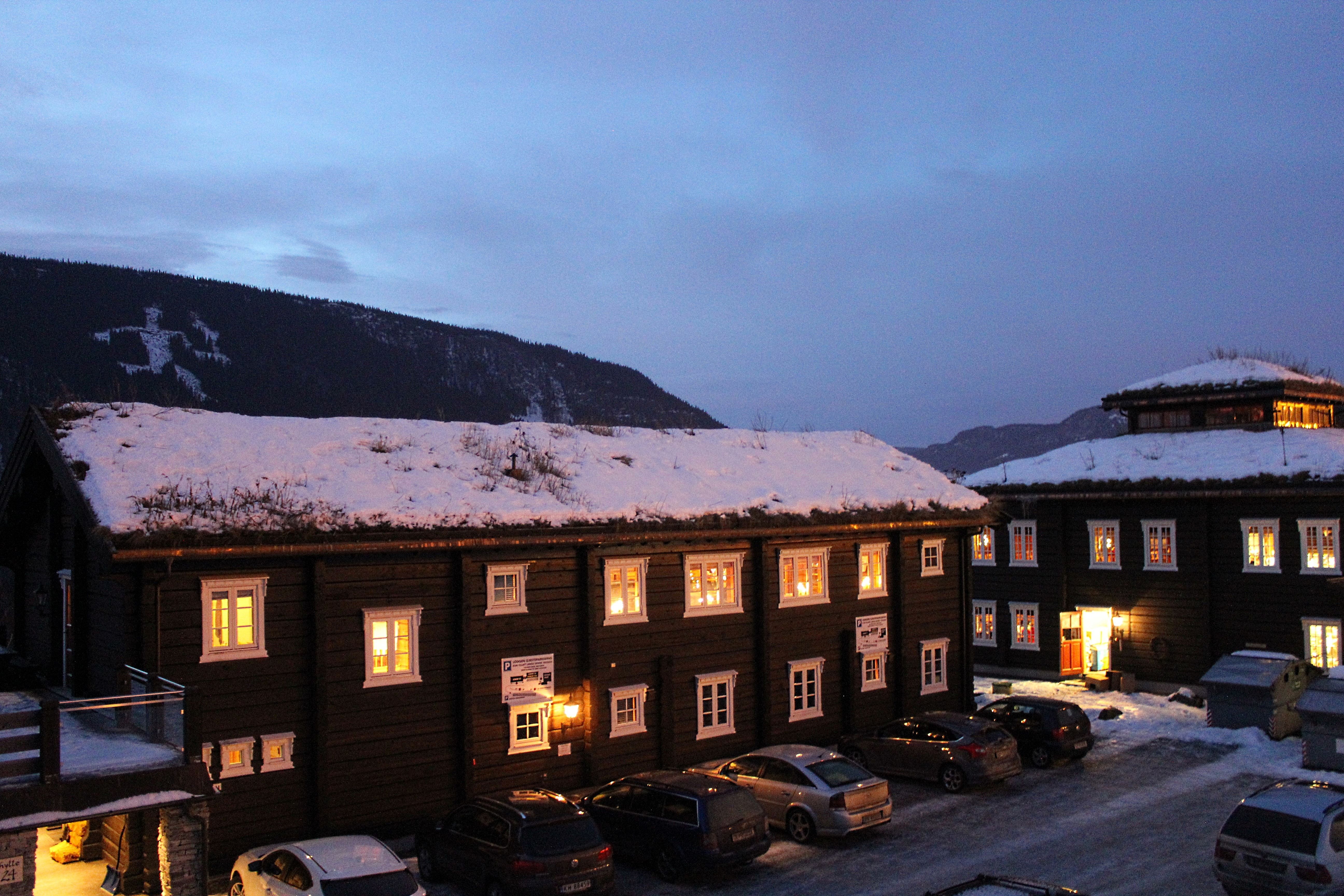 Snødekte trebygninger med opplyste vinduer ved en parkeringsplass, med fjell og blå kveldshimmel i bakgrunnen..