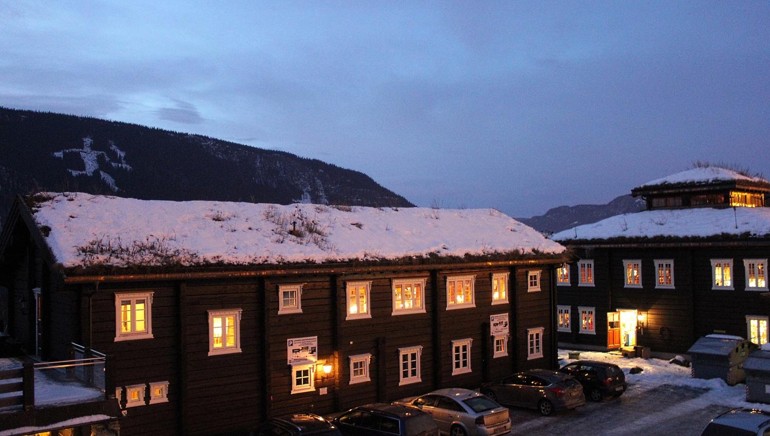 Snødekte trebygninger med opplyste vinduer ved en parkeringsplass, med fjell og blå kveldshimmel i bakgrunnen..