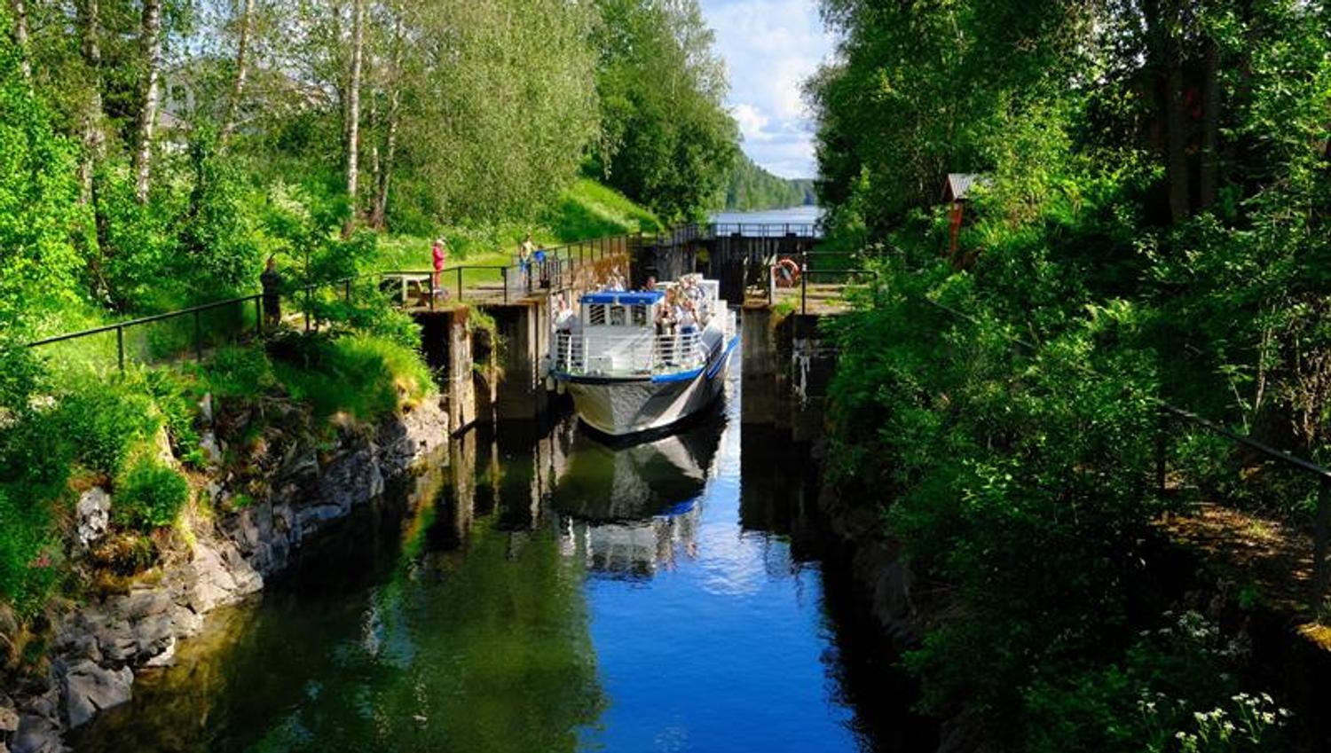 Gule blomster langs en elv ved en sluse, med frodig grønn vegetasjon på begge sider og en liten bro i bakgrunnen.