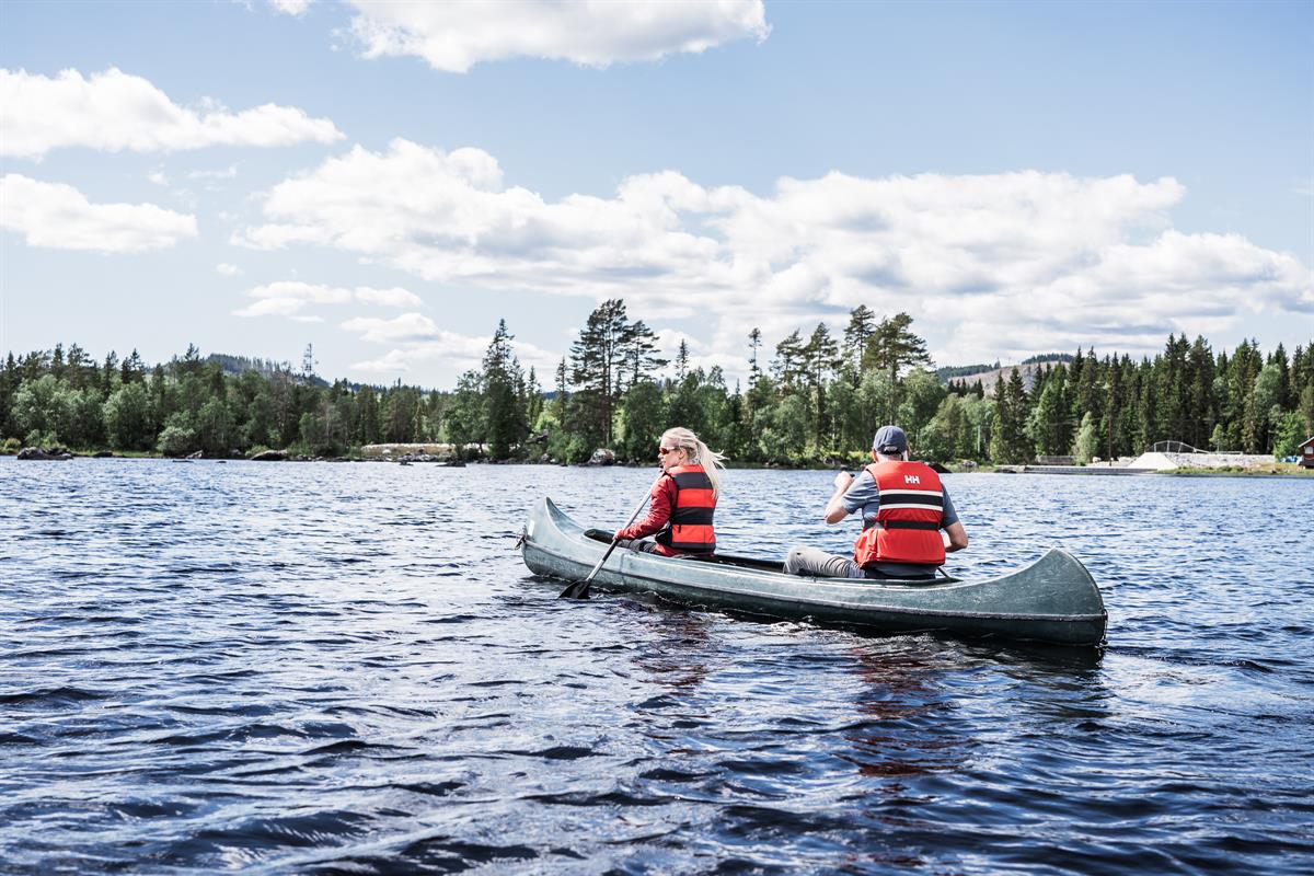 Two people paddling a canoe away from the camera on a lake, with a forested shoreline in the distance.