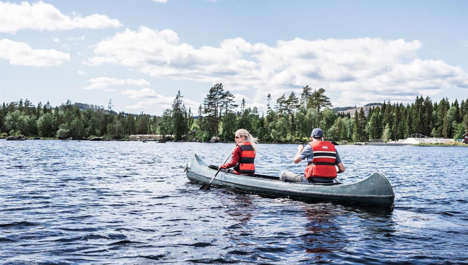 Two people paddling a canoe away from the camera on a lake, with a forested shoreline in the distance.