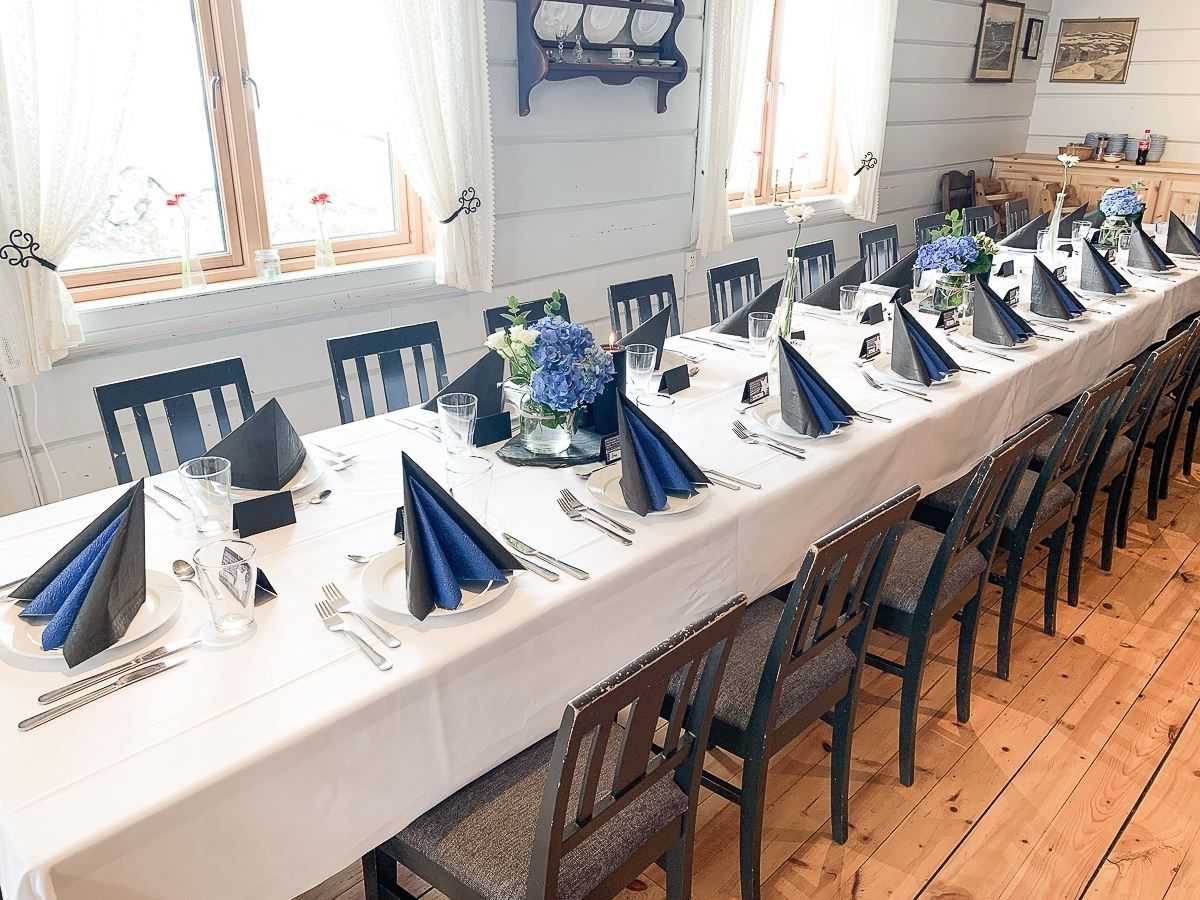 Long banquet table set with white cloth, folded dark napkins, glassware, and blue flower centerpieces by windows..