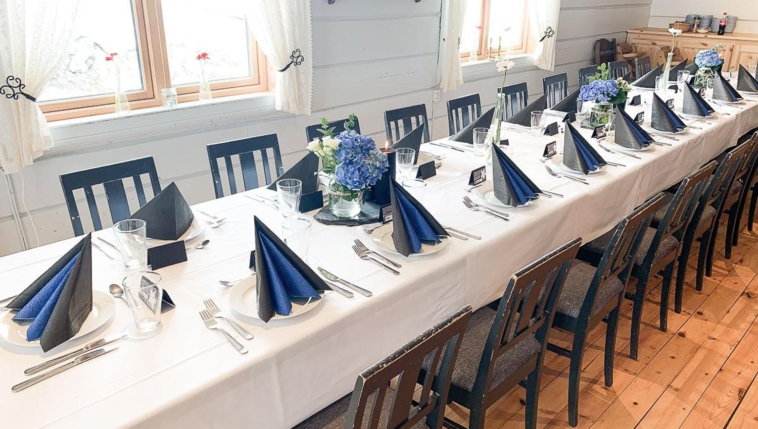 Long banquet table set with white cloth, folded dark napkins, glassware, and blue flower centerpieces by windows..