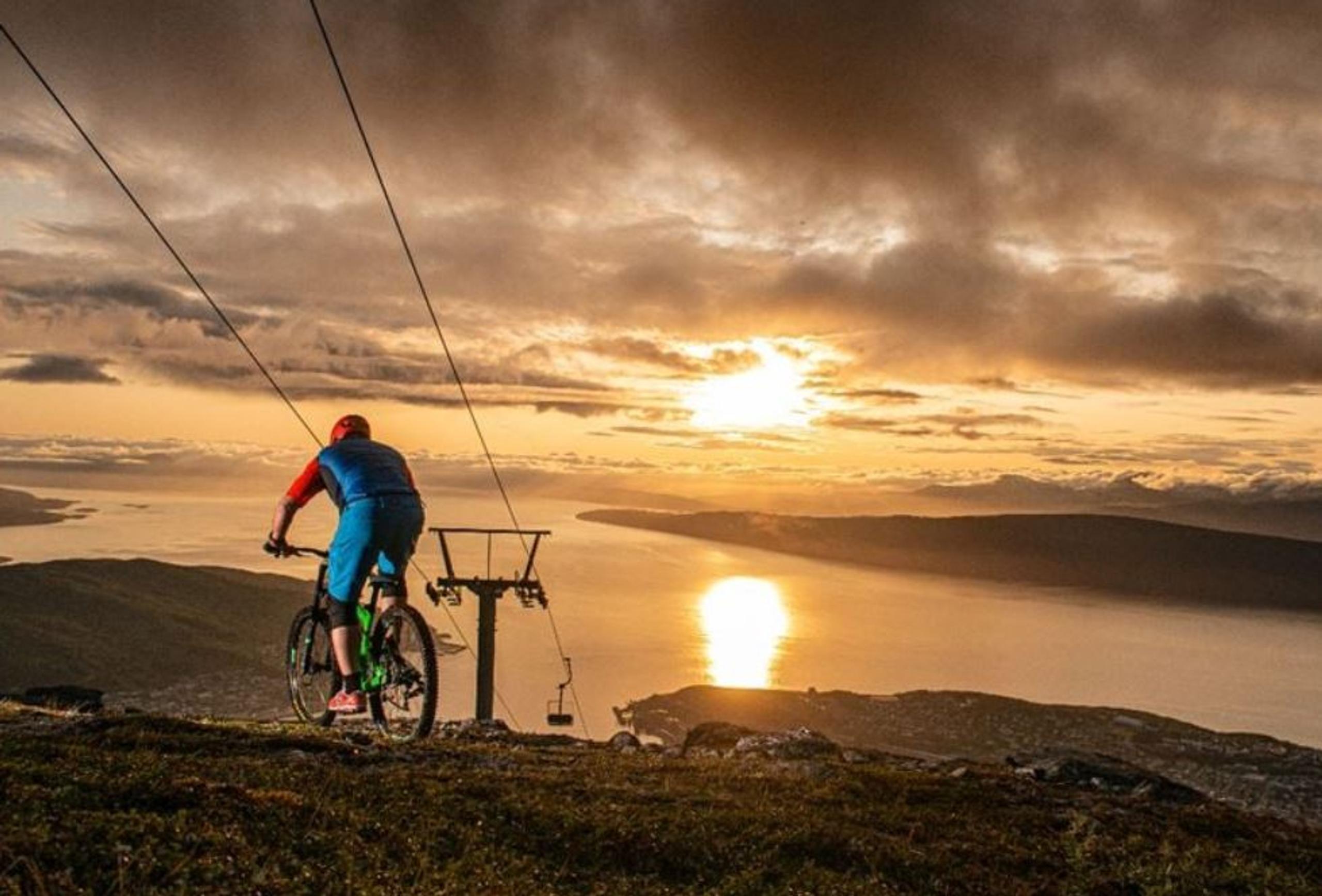 A cyclist in blue rides a mountain bike on a hillside near a lift line, overlooking a lake at sunset.