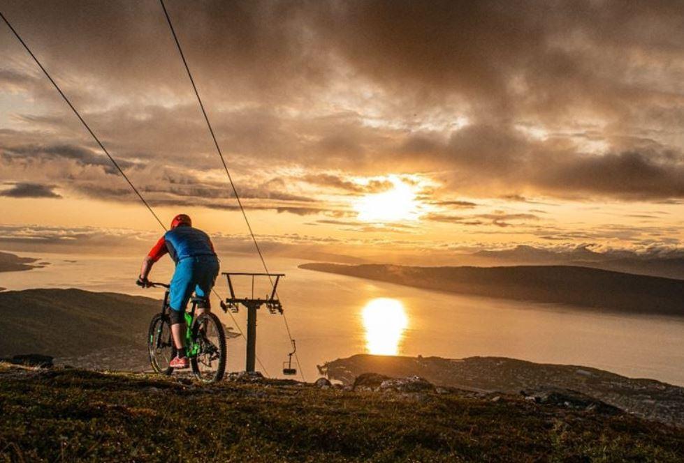 A cyclist in blue rides a mountain bike on a hillside near a lift line, overlooking a lake at sunset.