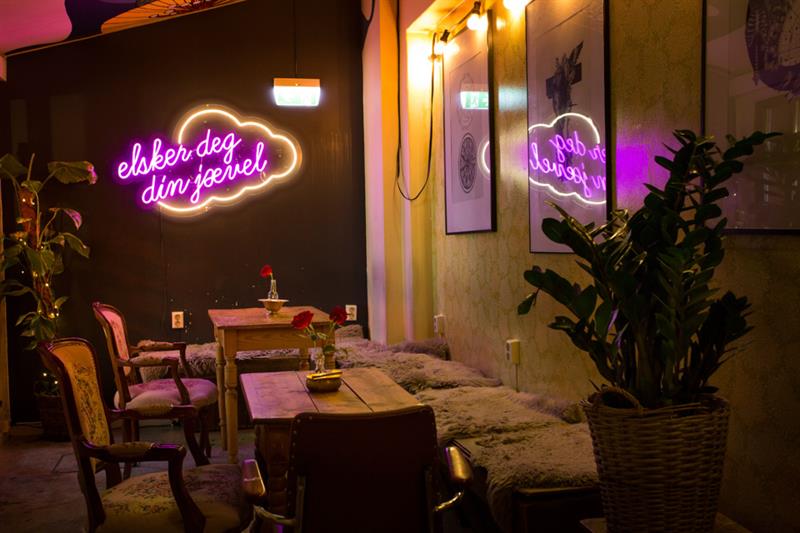 Interiør med bord, stoler og sitteseksjon med planter på hver side, samt neonskilt.