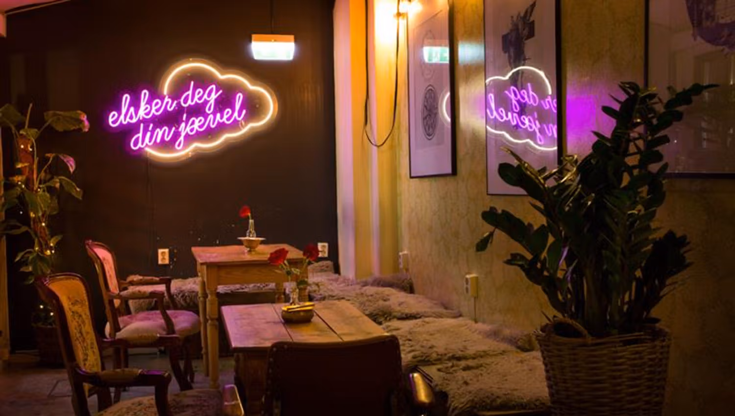 Interior with tables, chairs and a seating sections, as well as plants on each side, as well as a neon sign.