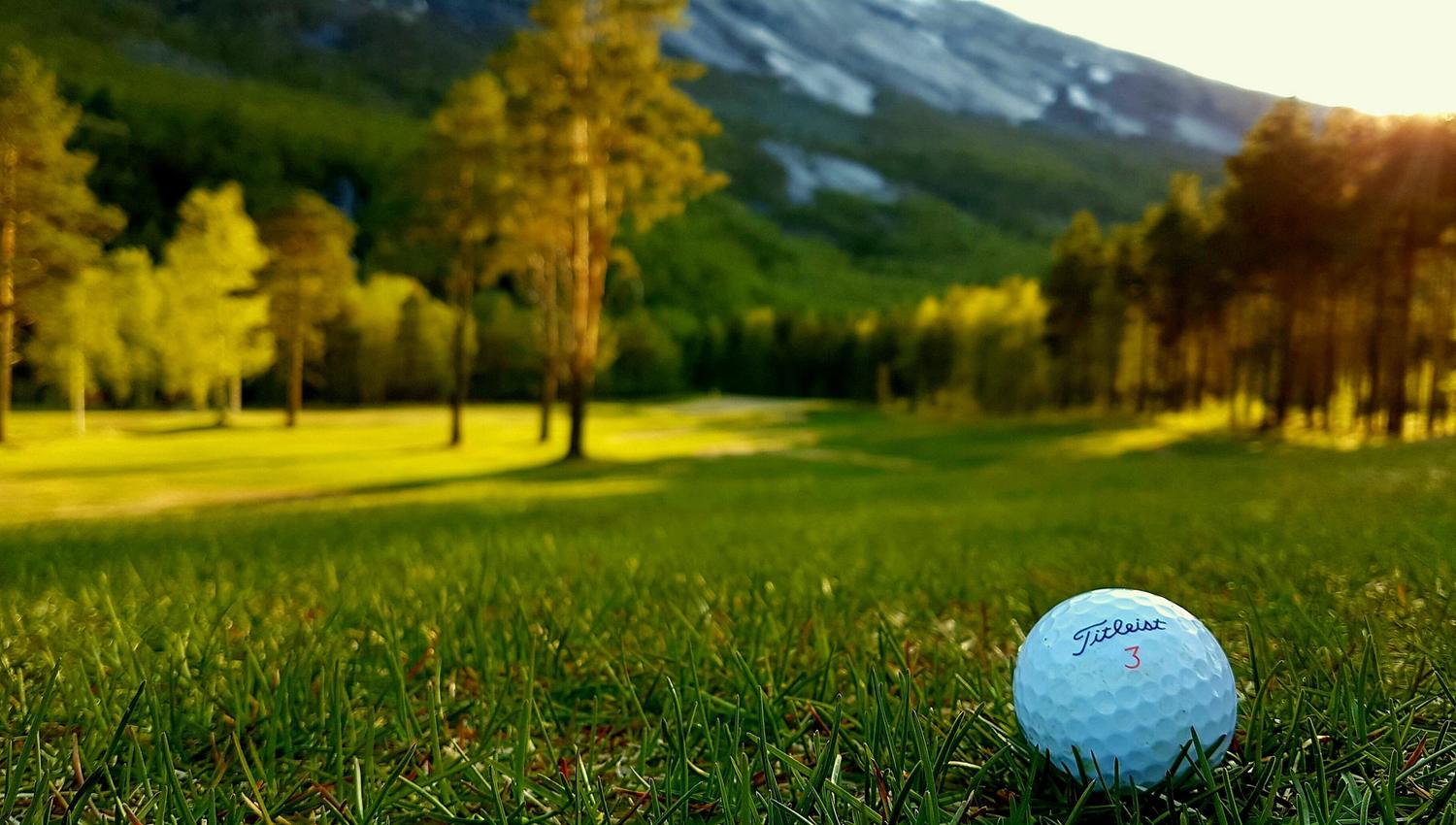 Close-up of a white golf ball in green grass with a sunlit fairway, trees, and a mountain in the background..
