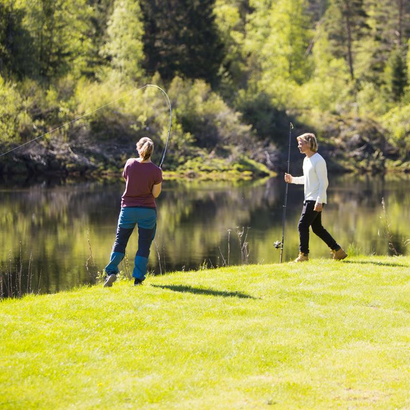 A couple standing on the grass by the river, fishing.