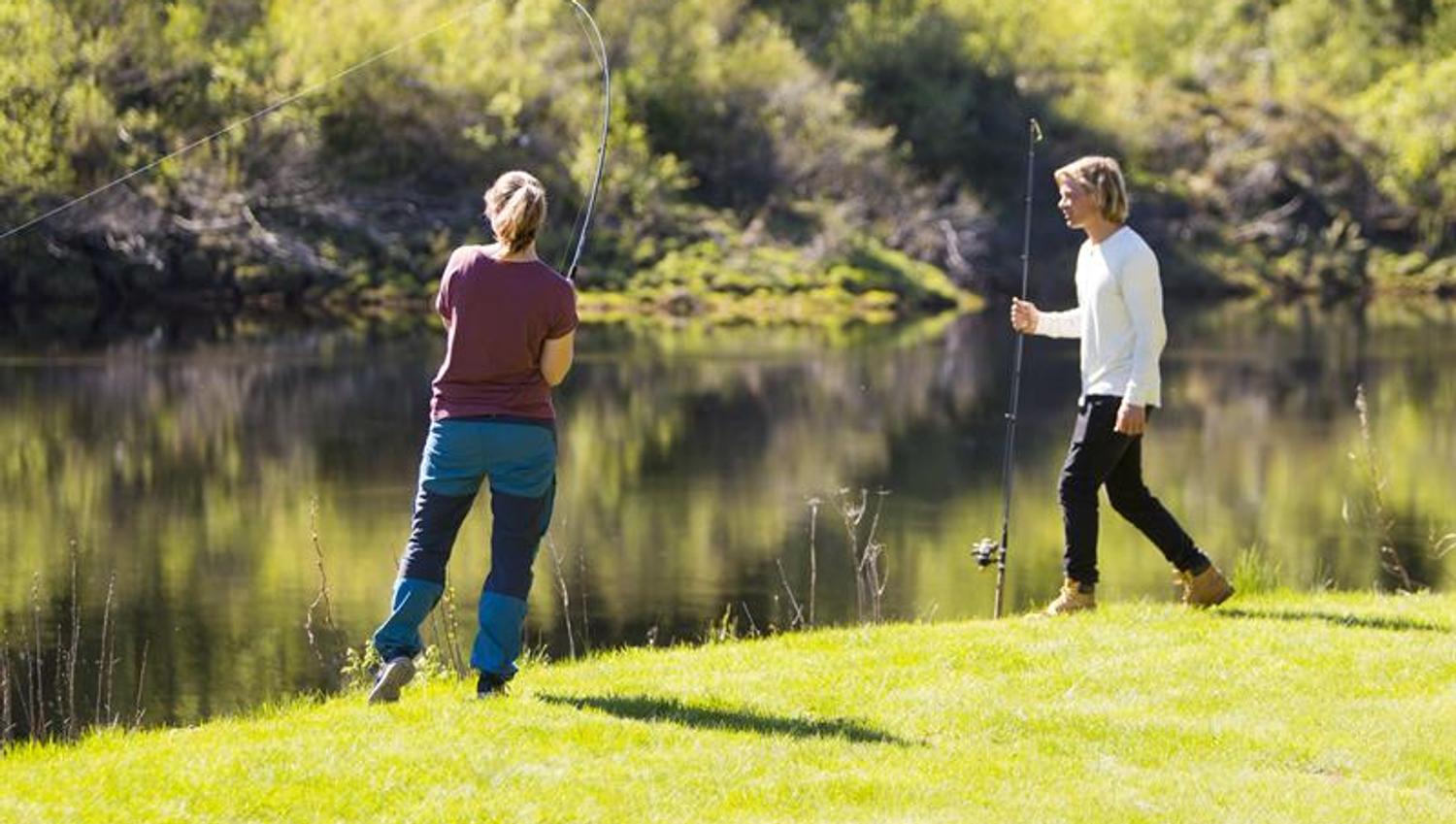 A couple standing on the grass by the river, fishing.