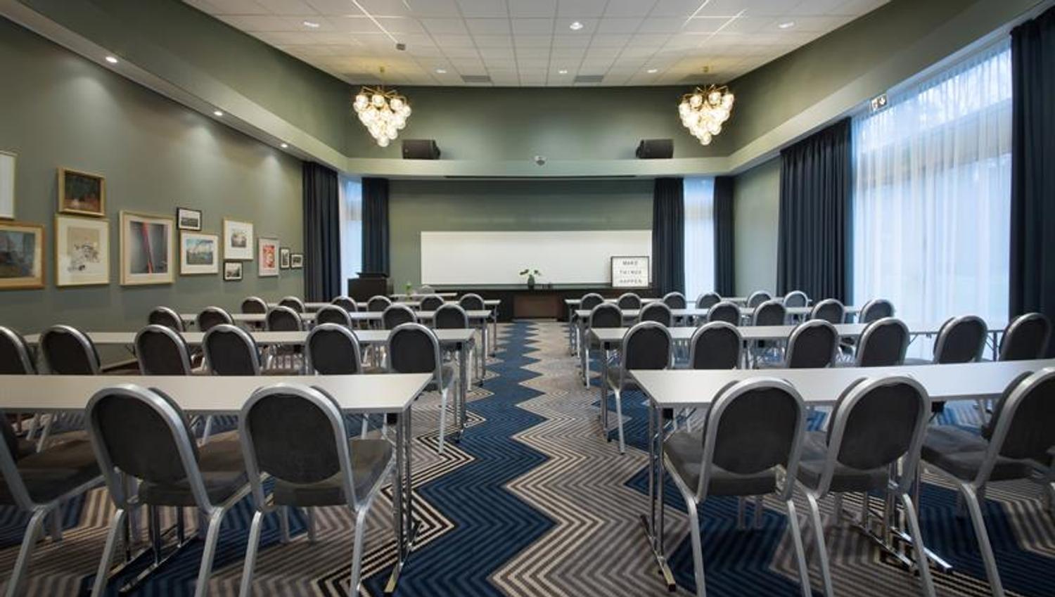 Konferanserom med bordoppsett i klasseromsstil, grå vegger og dekorative lysekroner i taket.