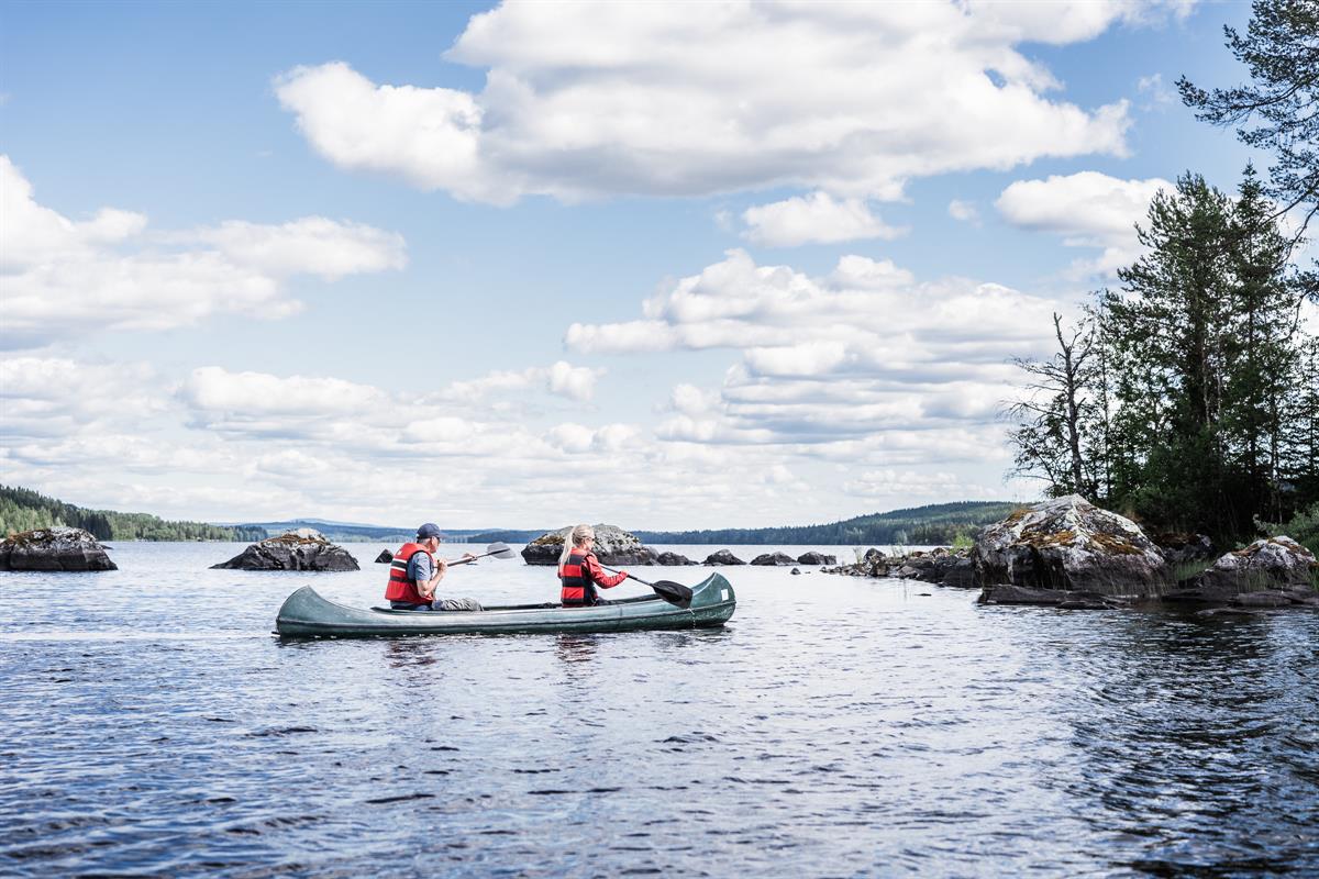 A wide lake landscape under a cloudy sky, with two people paddling a canoe near rocky islets.