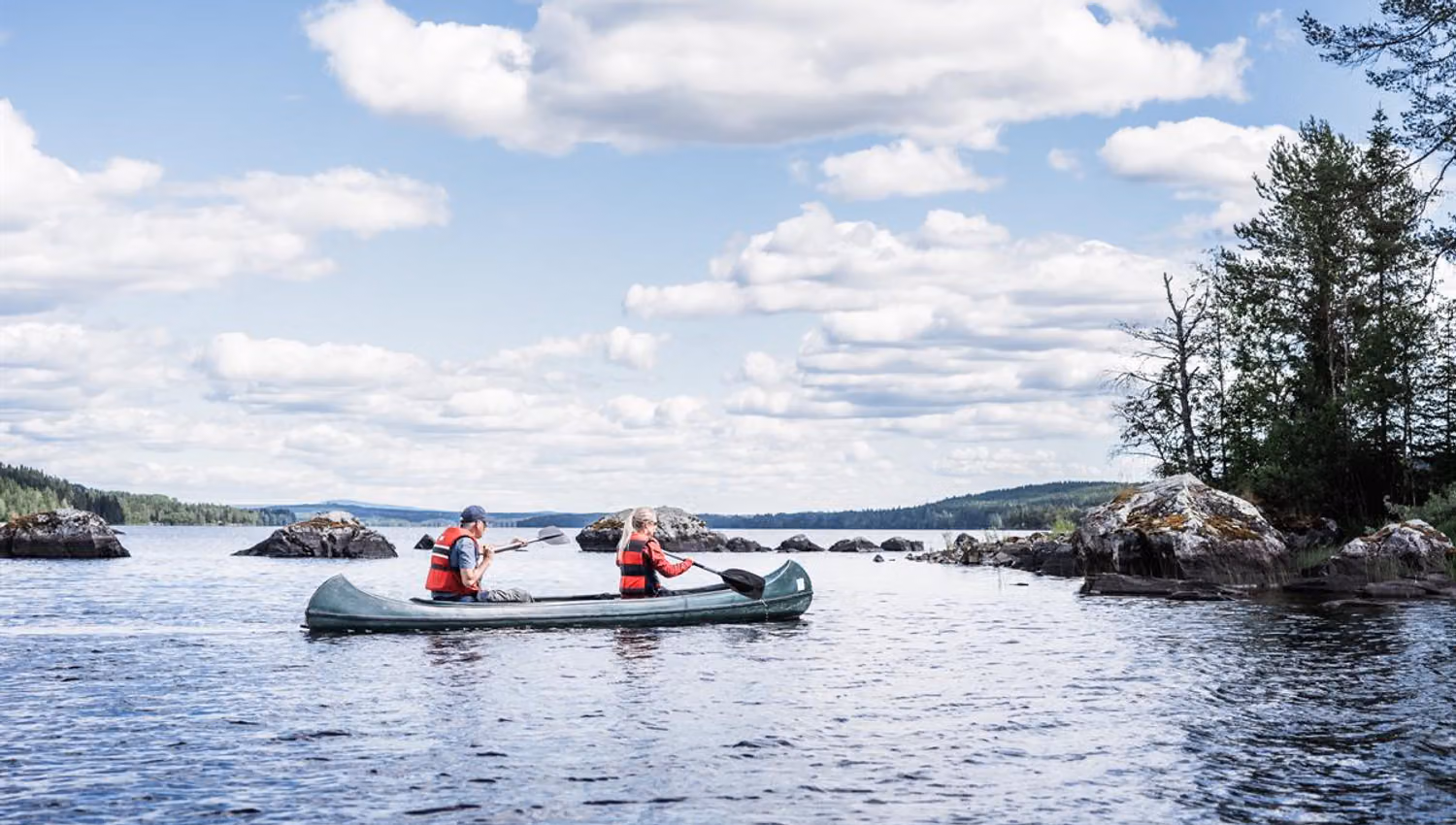 A wide lake landscape under a cloudy sky, with two people paddling a canoe near rocky islets.