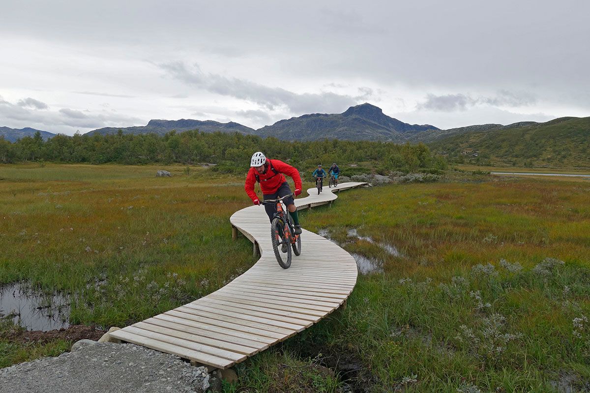 Cyclists on a sustainably built terrain cycling trail with wooden bridge elements over a swamp. Mountains in the background.