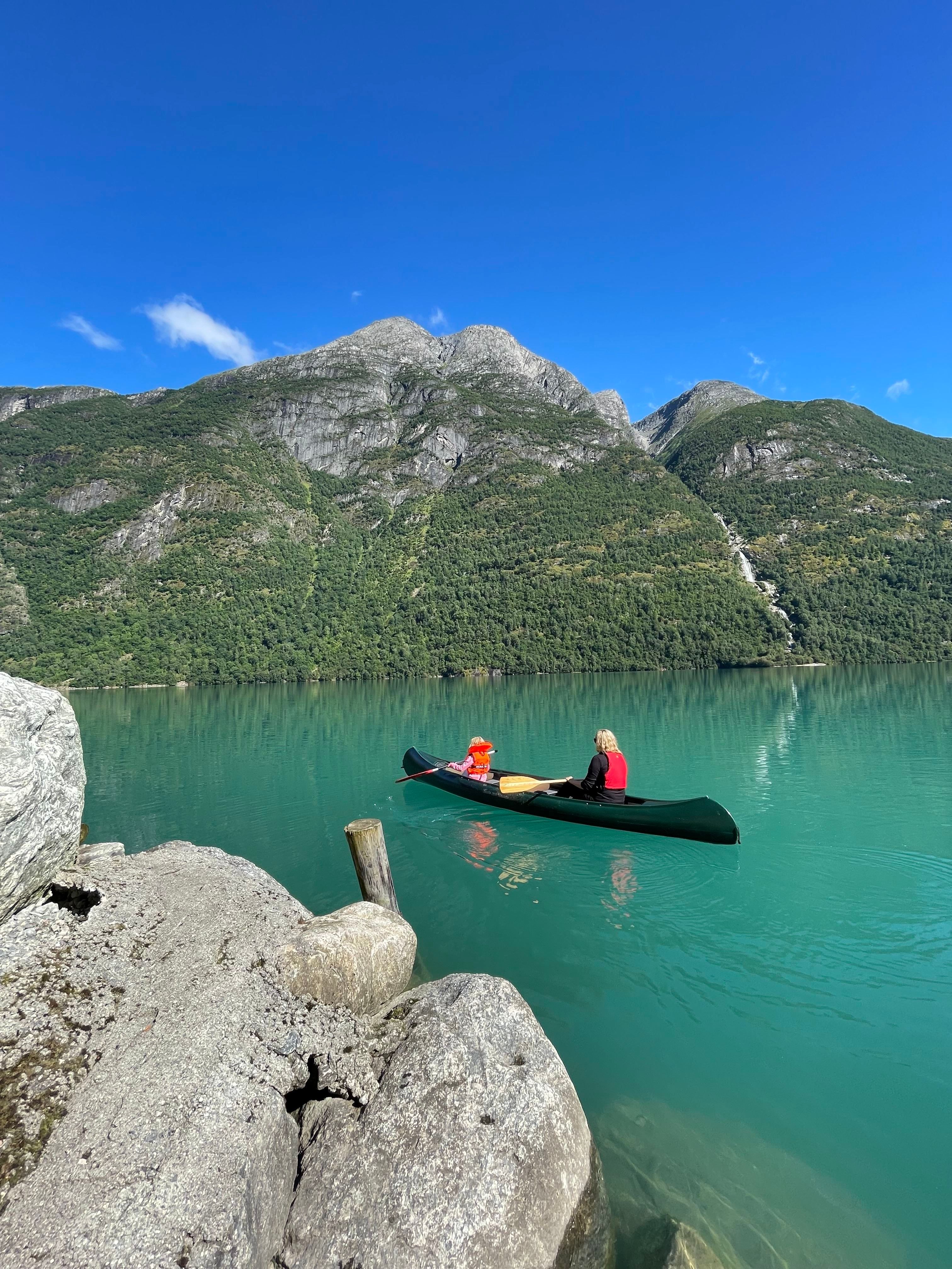 A person and a child in a canoe on turquoise lake water, with rocky shore and forested mountains under a blue sky..