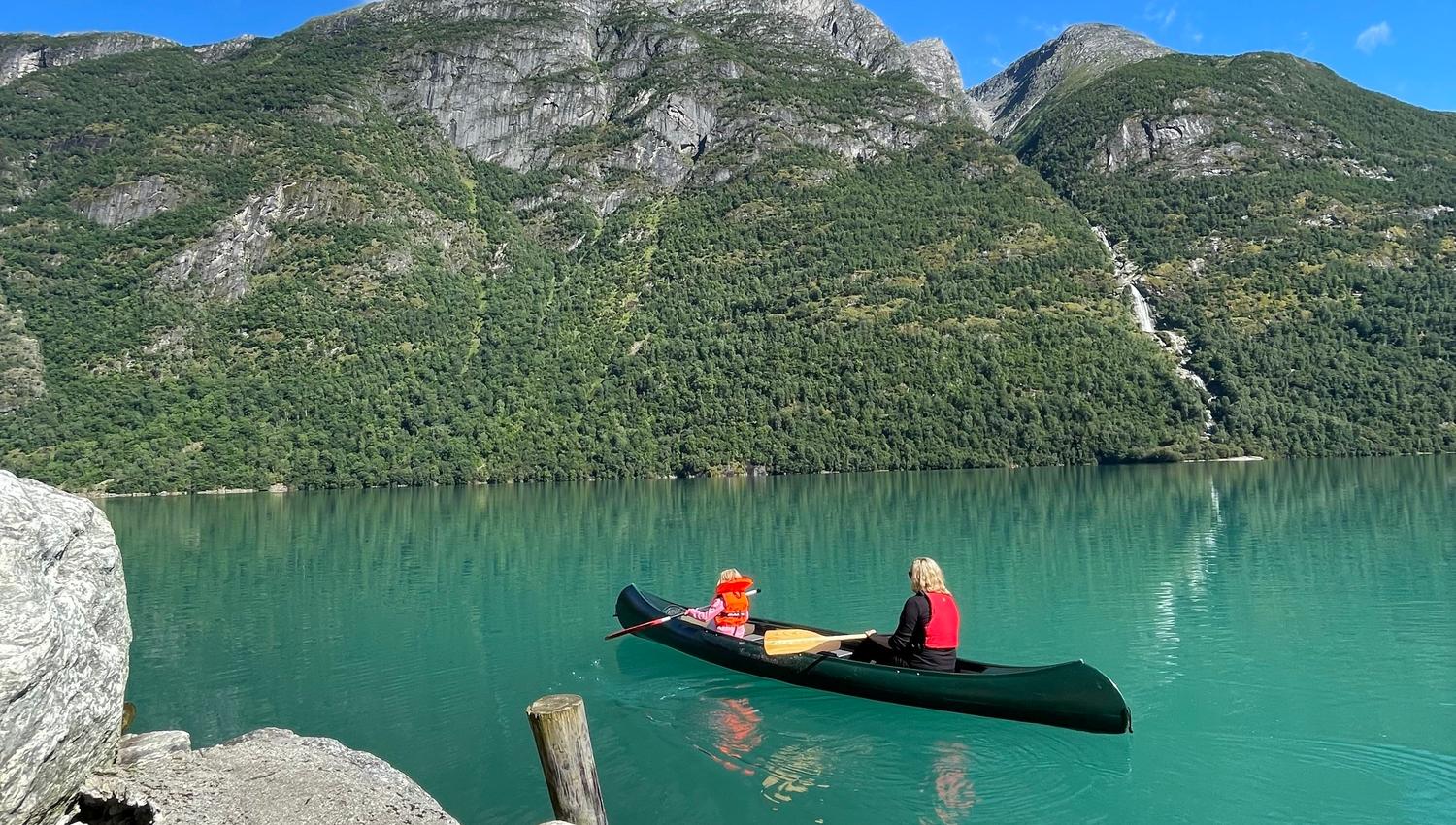 A person and a child in a canoe on turquoise lake water, with rocky shore and forested mountains under a blue sky..