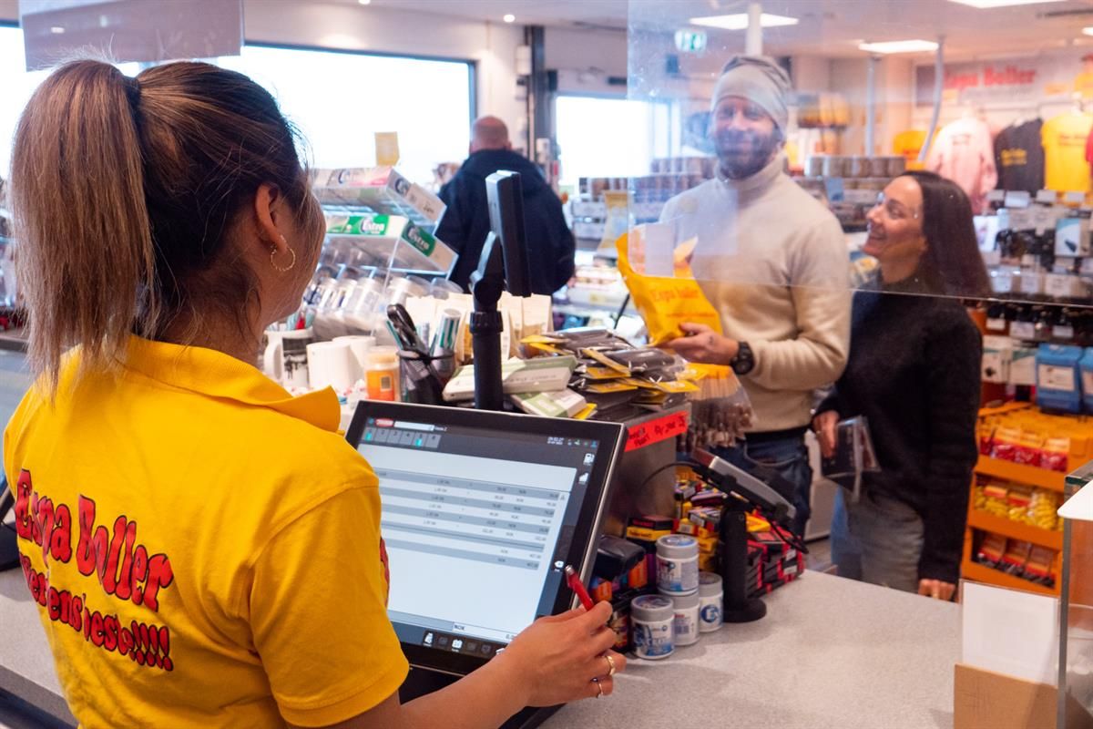 An employee in yellow workwear assists a customer at the checkout in a busy store, with food and beverages visible on the counter in a brightly lit re