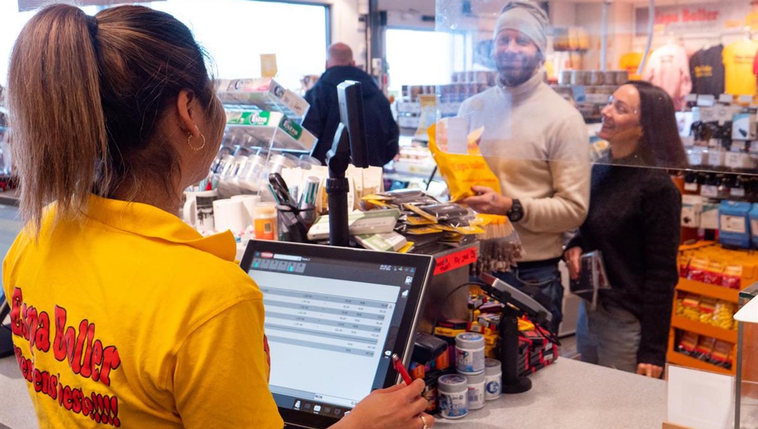 An employee in yellow workwear assists a customer at the checkout in a busy store, with food and beverages visible on the counter in a brightly lit re