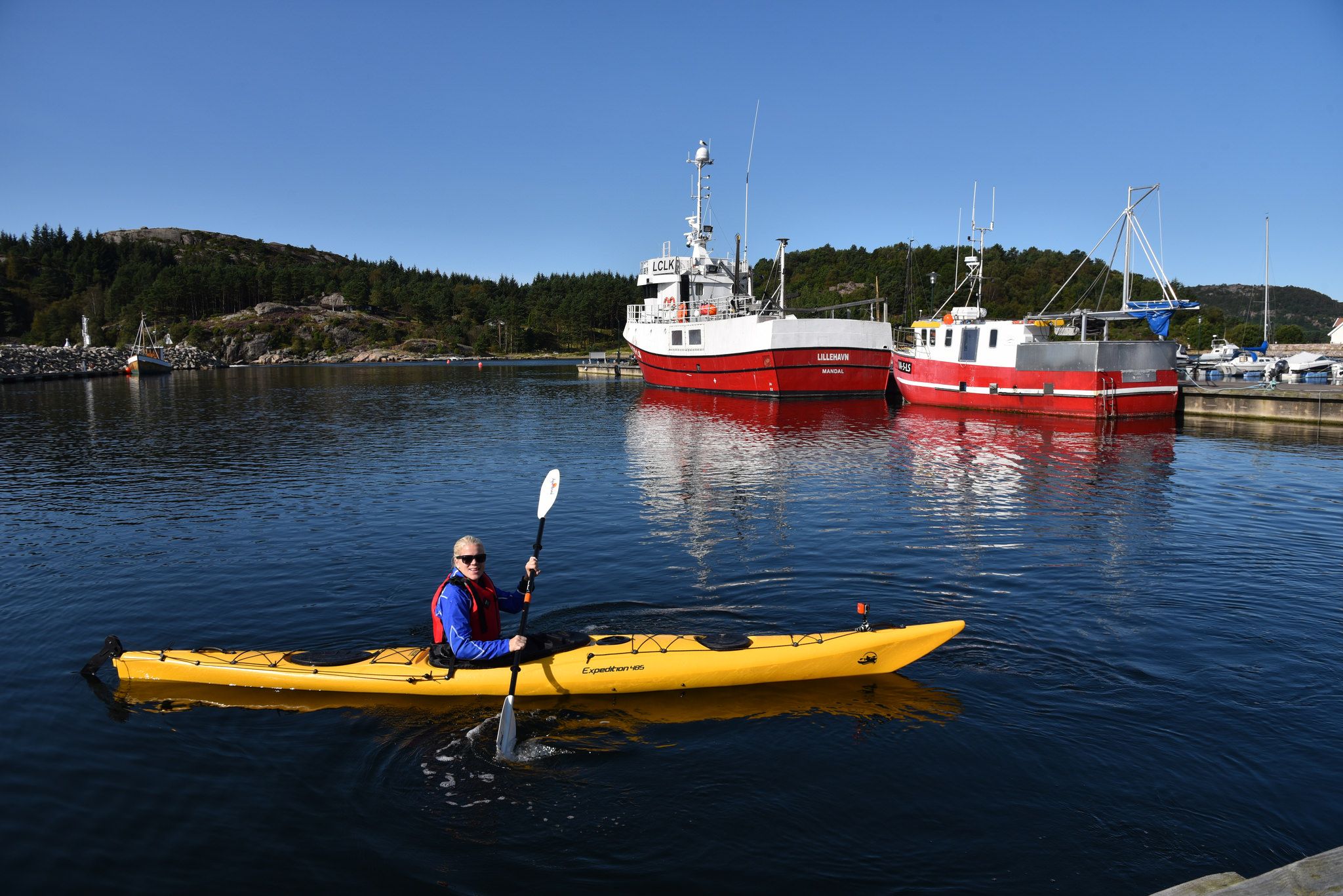 Kvinne i gul kajakk i vannet, med to store hvite og røde båter i bakgrunnen