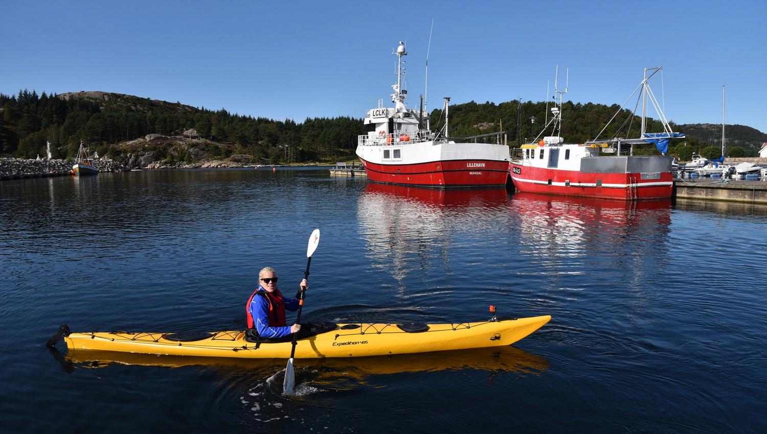 Kvinne i gul kajakk i vannet, med to store hvite og røde båter i bakgrunnen