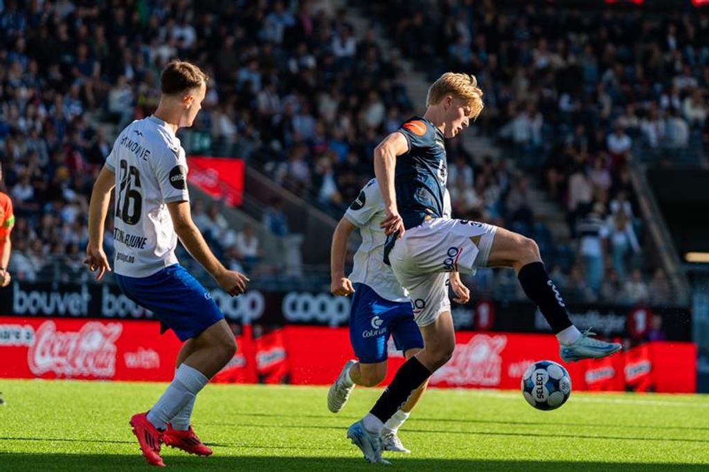 En fotballspiller spiller ballen mellom to motspillere foran fullsatte tribuner