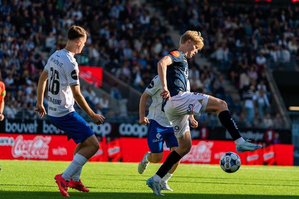 A footballer dribbles the ball between two opponents in front of a packed crowd