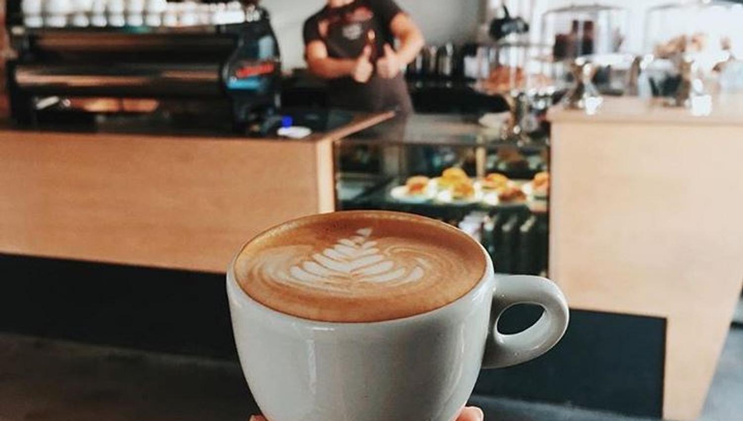 en hånd som viser frem en kaffe med kaffebaren i bakgrunnen