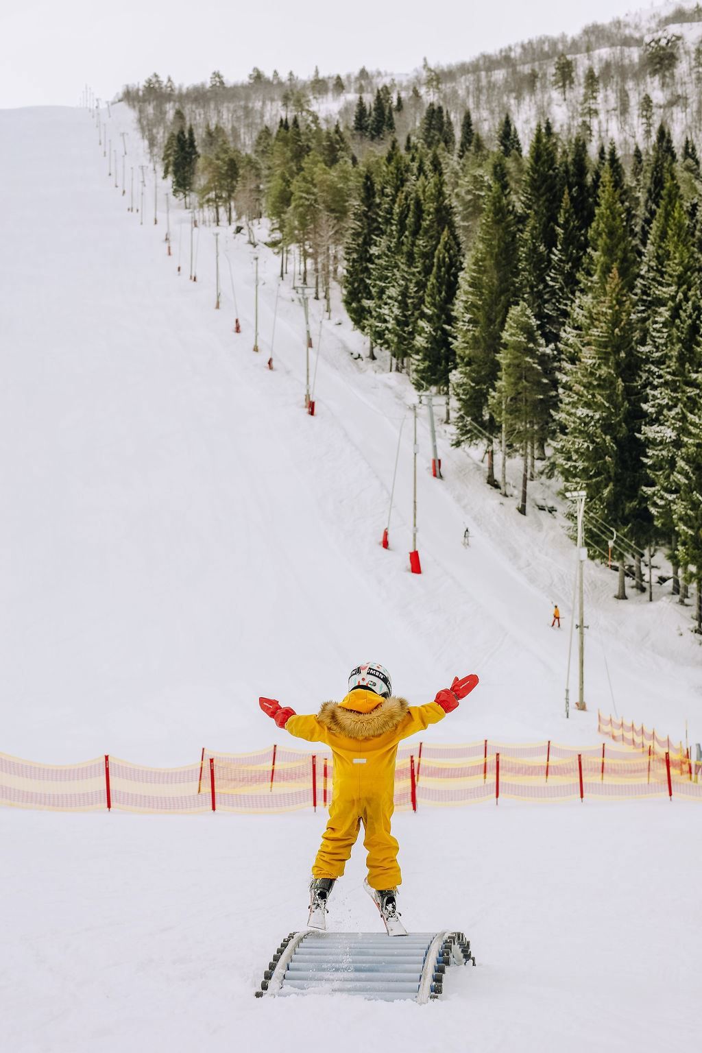 Child in a yellow snowsuit stands on skis with arms raised on a snowy slope beside pine trees and fencing.