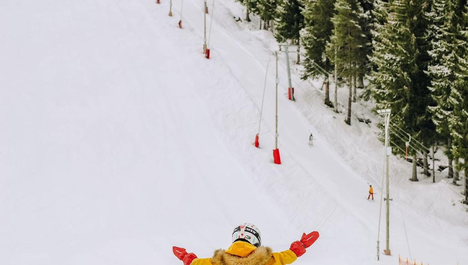 Child in a yellow snowsuit stands on skis with arms raised on a snowy slope beside pine trees and fencing.