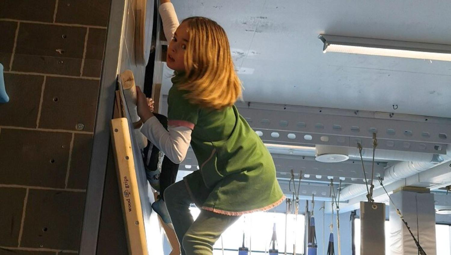 A child climbs an indoor climbing wall in a gym, reaching for a wooden hold with hanging obstacles behind..
