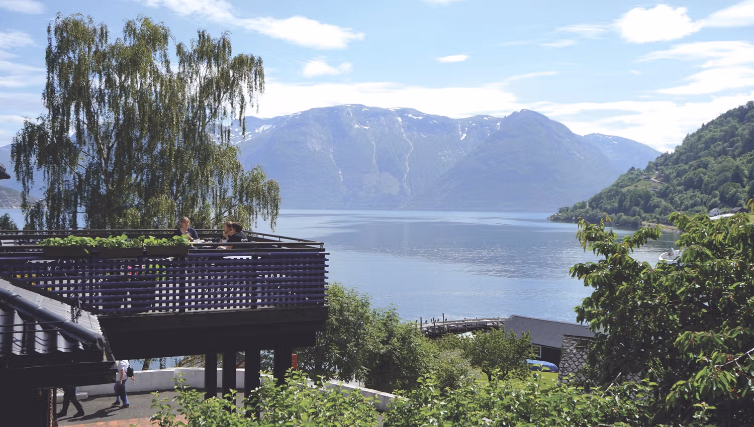 Utsikt ut over fjord og fjell. I forgrunnen er det grønne trær og en altan hvor det sitter tre mennesker.