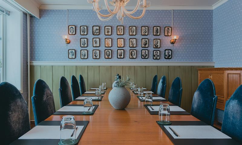 A meeting room with a table, blue chairs and walls. with a collection of old portraits on a wall in the background