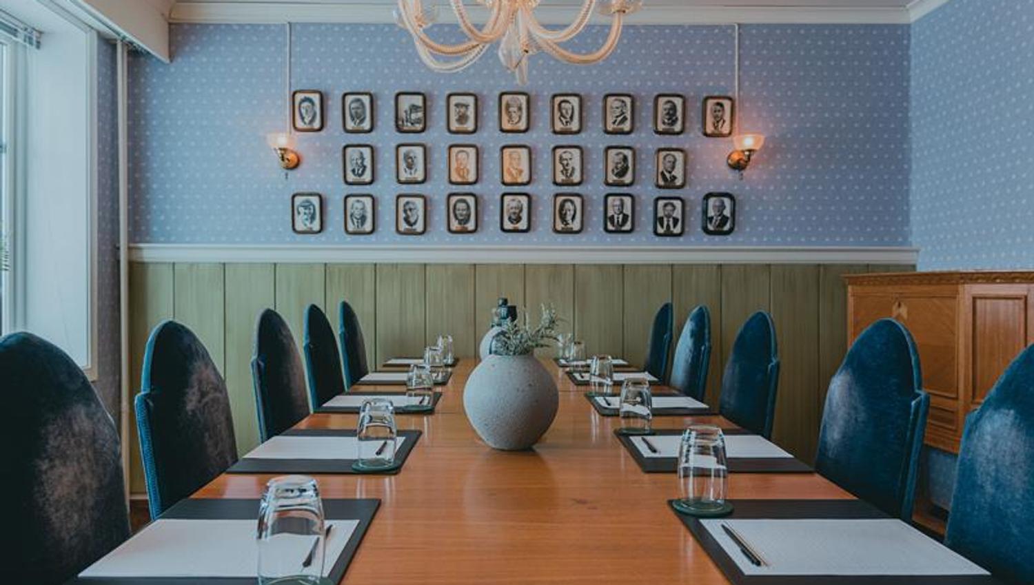 A meeting room with a table, blue chairs and walls. with a collection of old portraits on a wall in the background