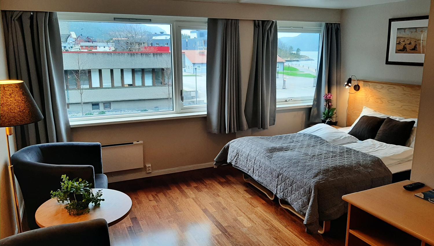 Lyst hotellrom med dobbeltseng, lenestolar, lite bord og store vindauge med utsyn mot bygningar og sjø..