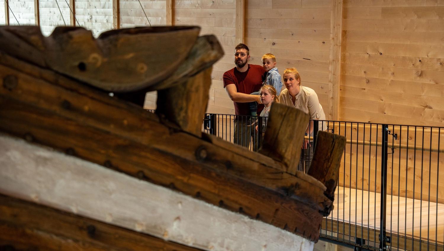 A small group stands behind a railing in a wooden interior, looking toward a large rustic wooden object in front..