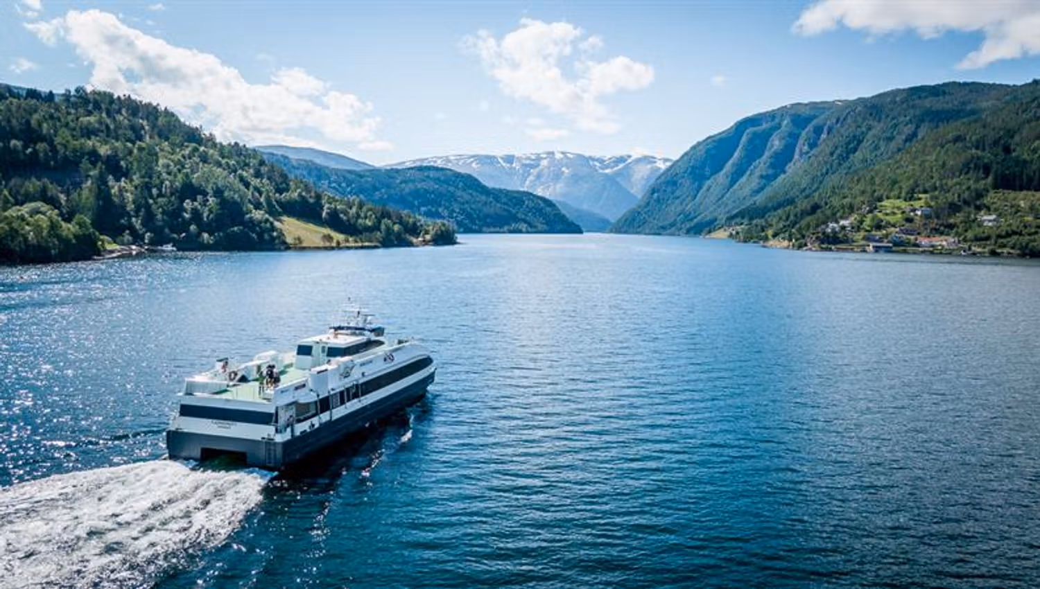 Fjordcruisebåten Fjorddrott seiler innover Hardangerfjorden med snødekte fjell i bakgrunnen, sett frå drone.