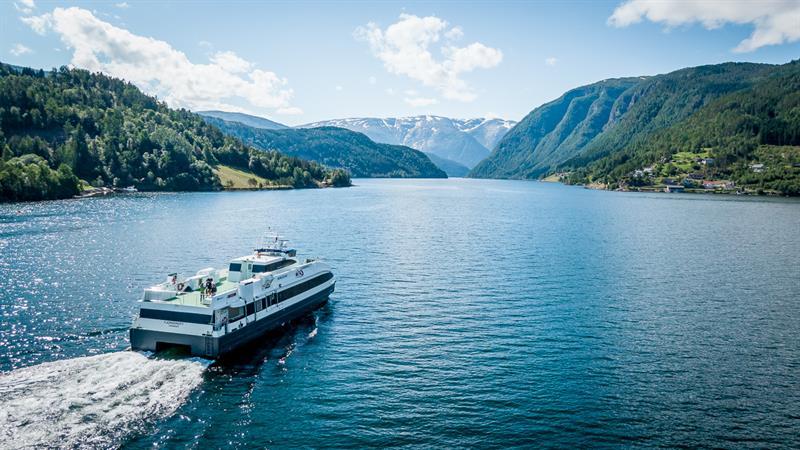Fjordcruisebåten Fjorddrott seiler innover Hardangerfjorden med snødekte fjell i bakgrunnen, sett frå drone.