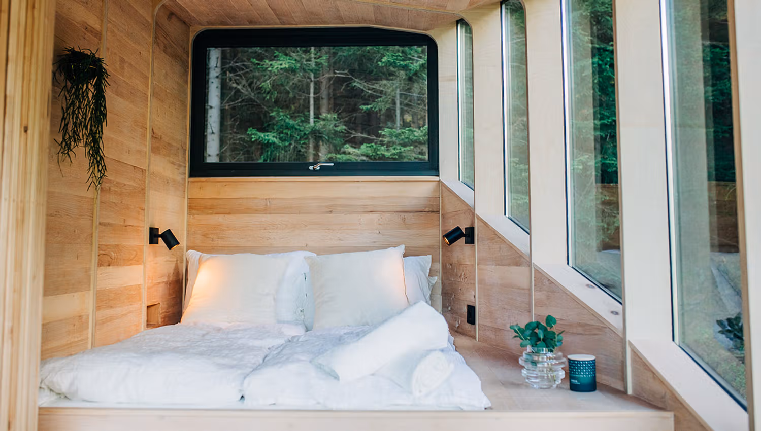 Ei seng med mjuke tekstilar og store vindauge som slepp inn naturlyset i Woodnest tretopphytte.