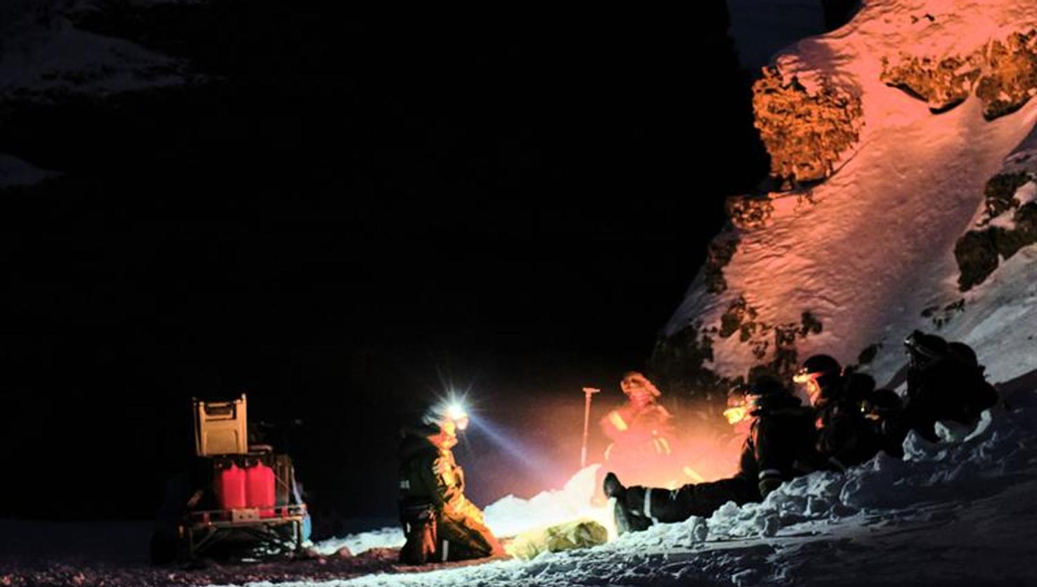 People are sitting on the ground next to a stony cliff. Mountian in the background. Clear starsky.