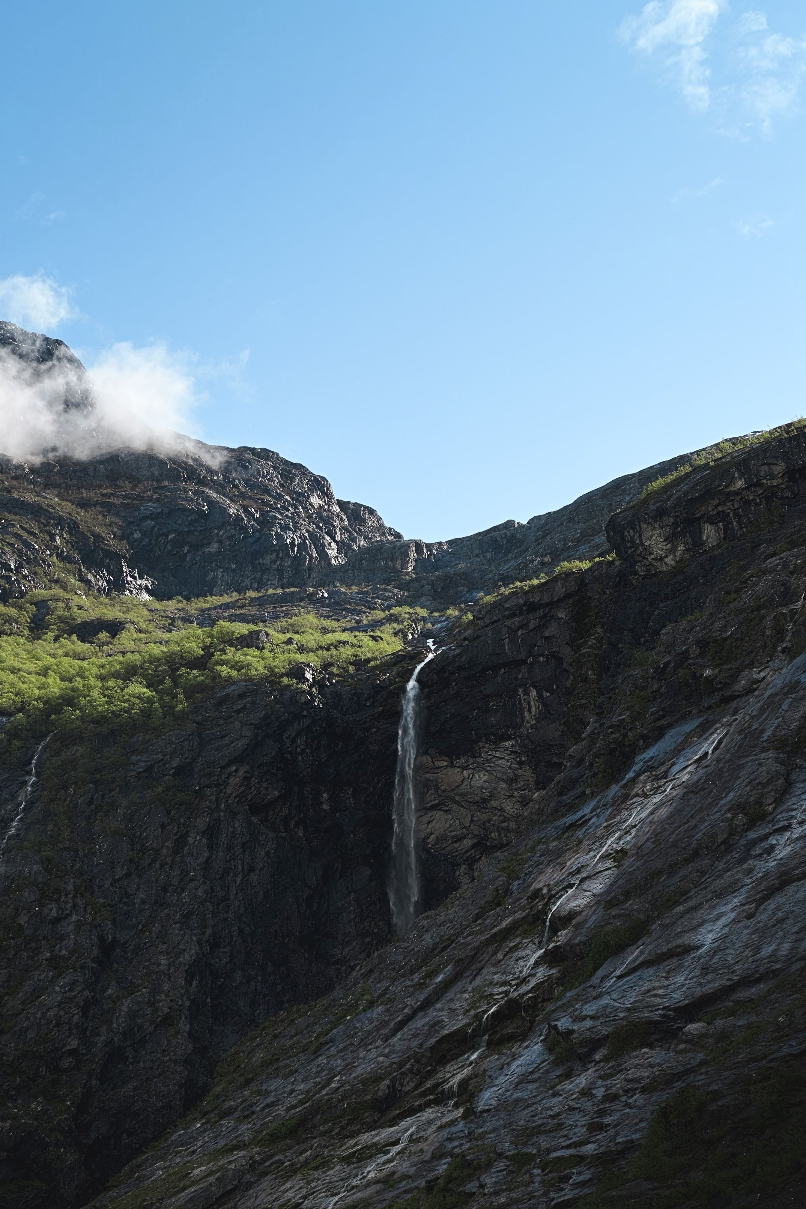 A narrow waterfall drops down dark rocky cliffs with green shrubs below a bright blue sky with light clouds..