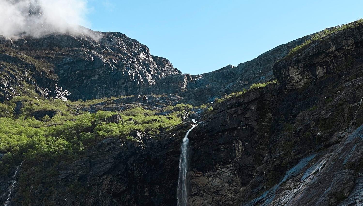 A narrow waterfall drops down dark rocky cliffs with green shrubs below a bright blue sky with light clouds..