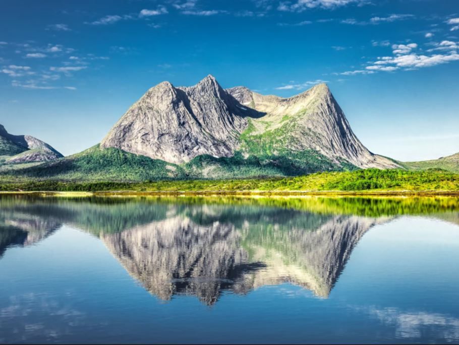 Ruggete fjell med grønn vegetasjon speiles tydelig i en stille innsjø under blå himmel med skyer..