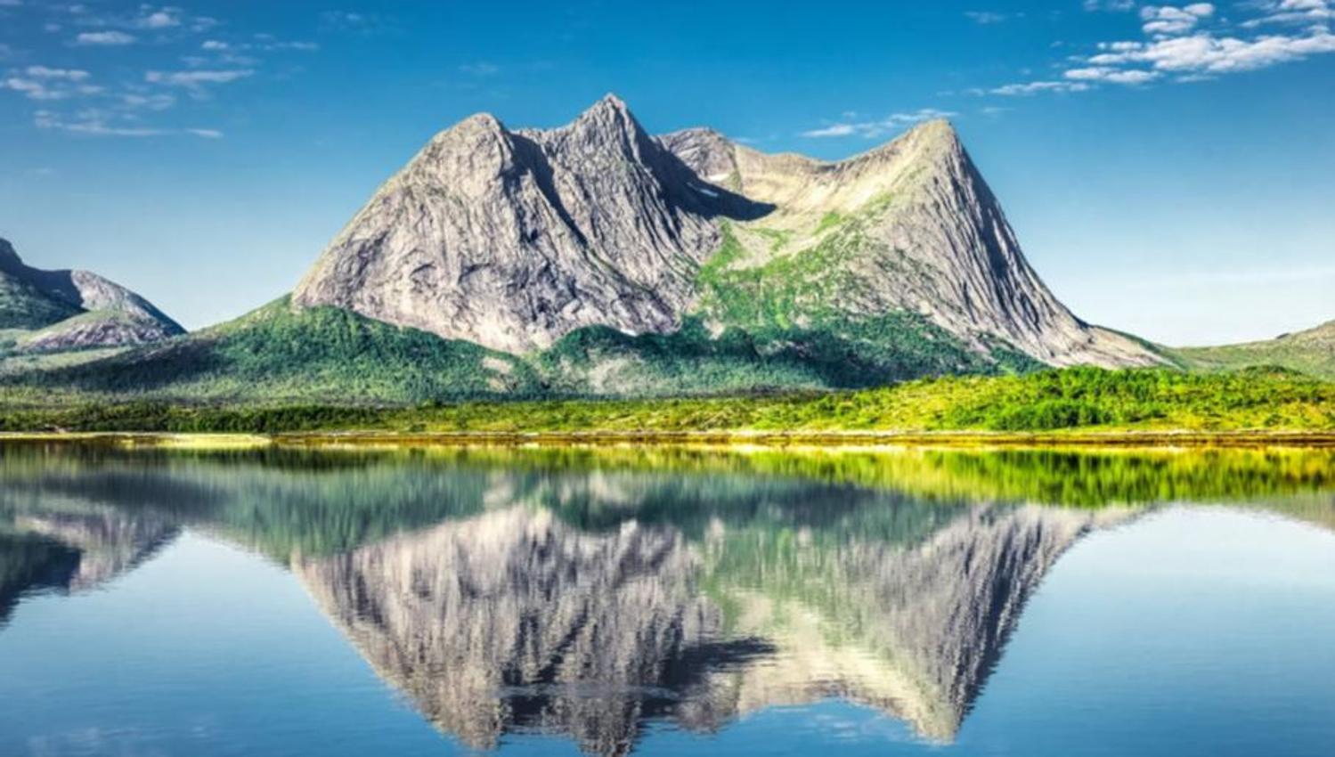 Ruggete fjell med grønn vegetasjon speiles tydelig i en stille innsjø under blå himmel med skyer..