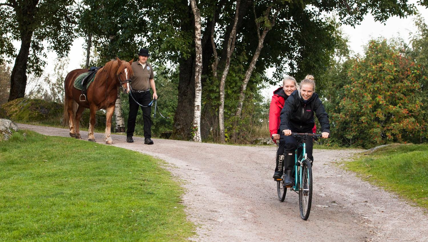 Two smiling girls are riding a tandem bicycle, and a man is leading a brown horse with a white blaze behind the girls.