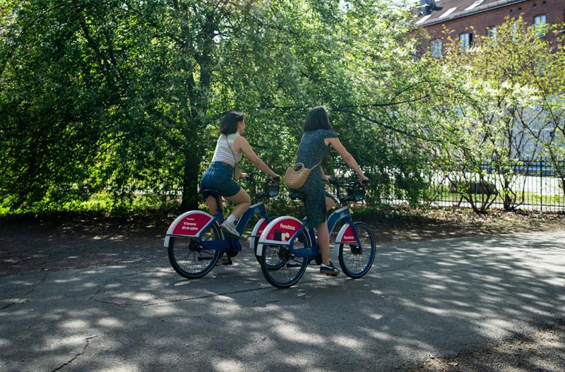 To jenter som sykler langs en vei, med et tre og et hus i bakgrunnen