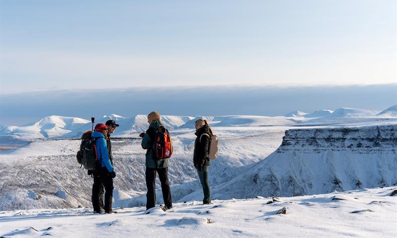 flere personer står på toppen av snødekt fjell. i bakgrunnen ses flere platåer og snødekte fjell