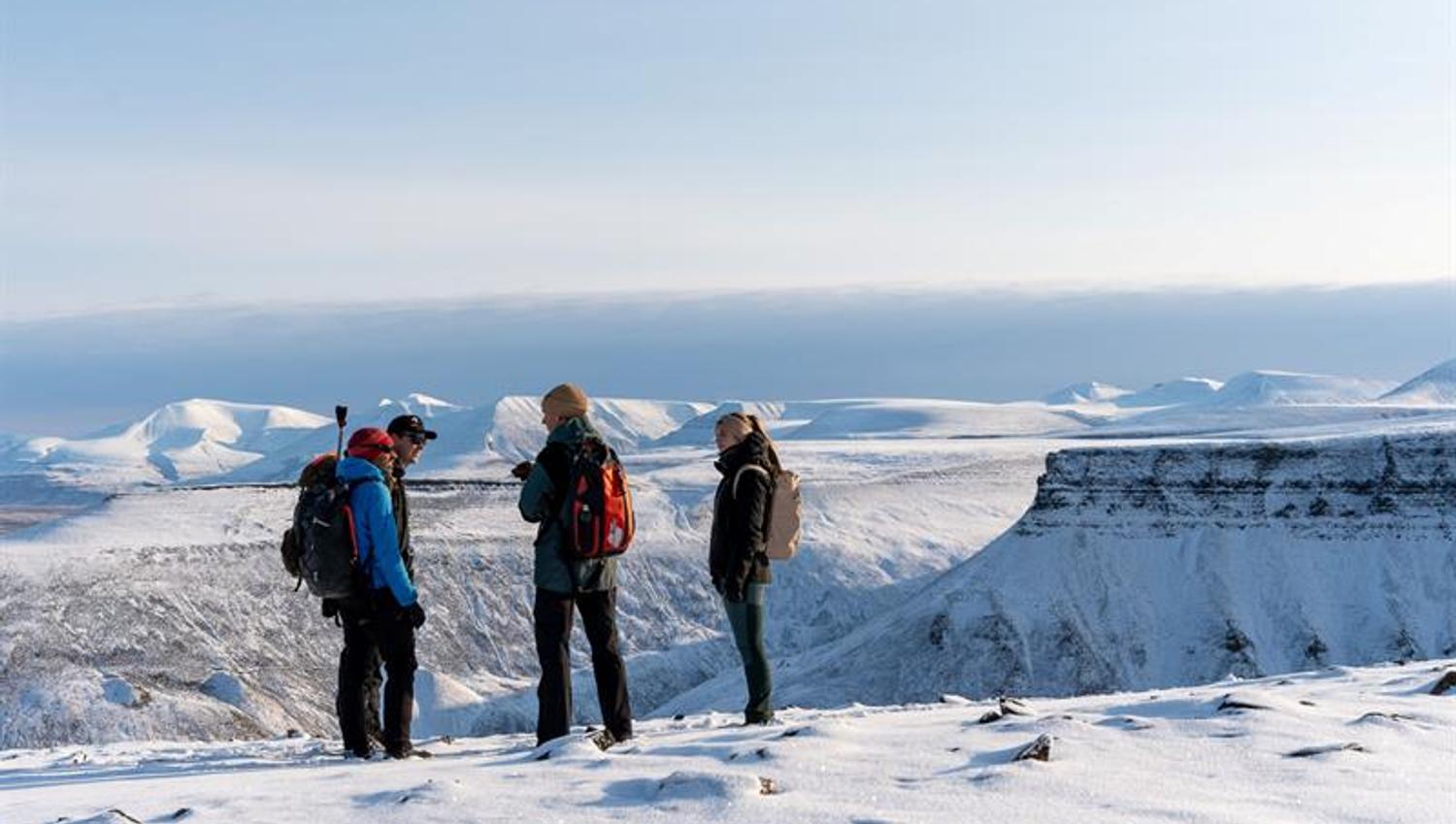flere personer står på toppen av snødekt fjell. i bakgrunnen ses flere platåer og snødekte fjell