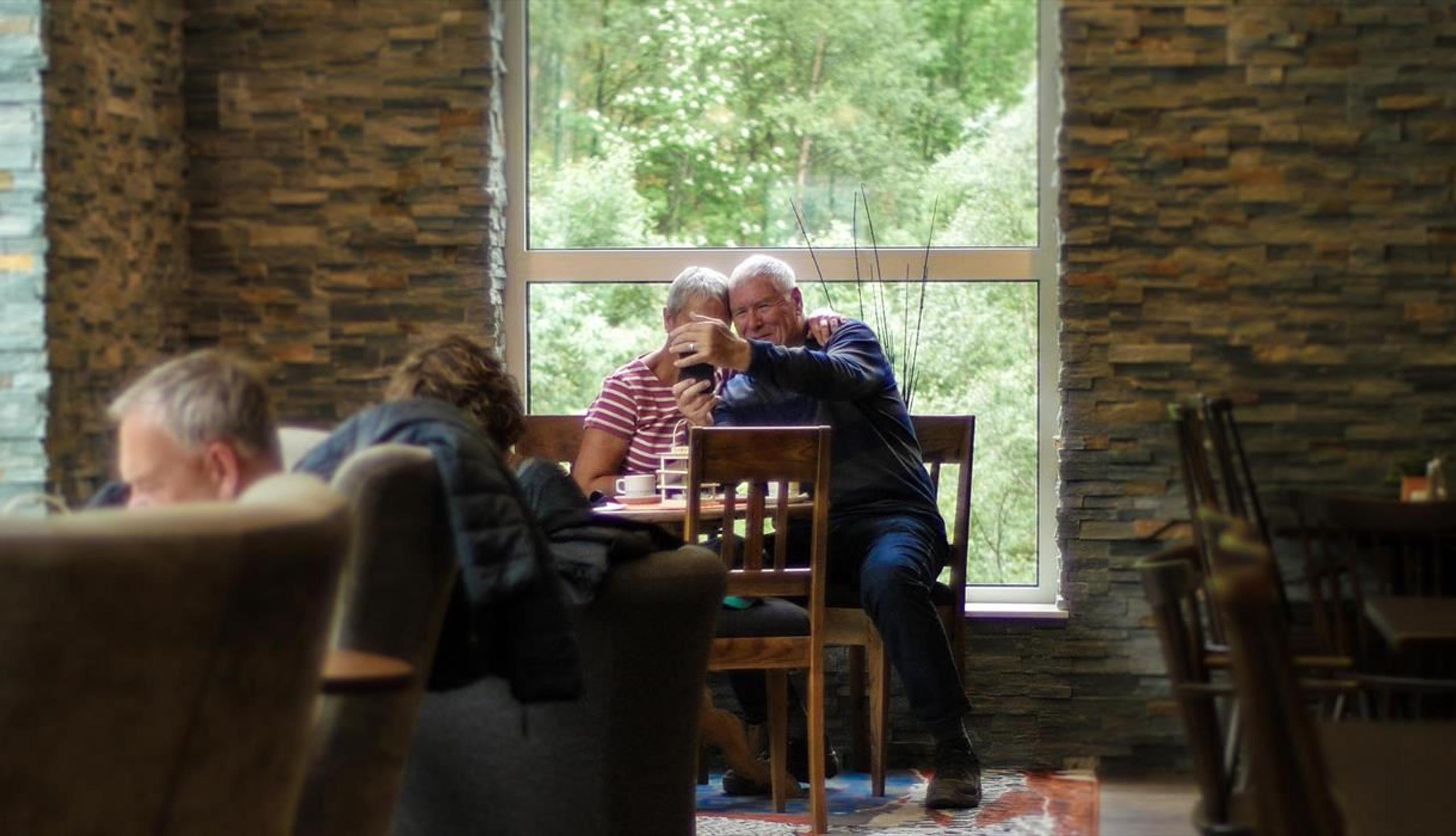 Eit eldre par sit ved eit bord i ein kafé og tek ein selfie, med stort vindauge og grøne tre i bakgrunnen..